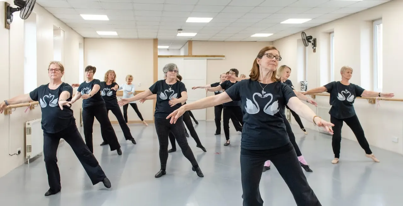 Group of adults in a dance studio wearing matching black t-shirts with swan design, performing a synchronized dance routine at the barre.