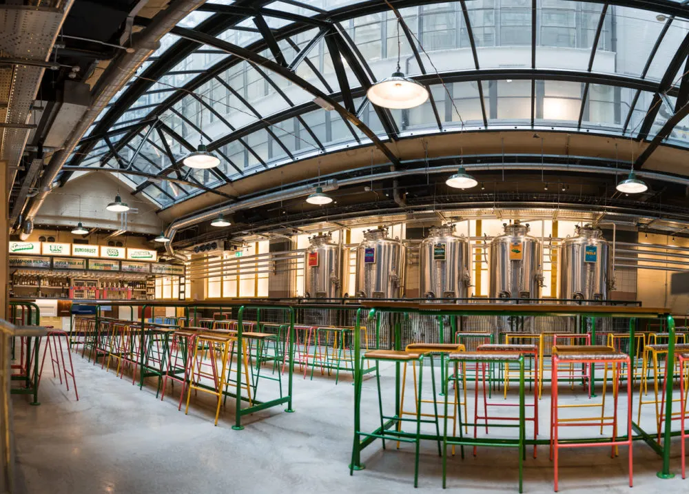 Interior of a brewery or distillery with colourful railings, seating areas, and large stainless steel vats under an arched glass roof.