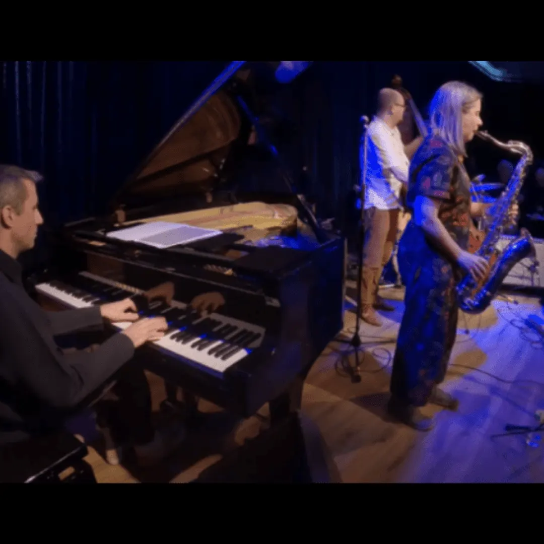 Jazz band performing on stage with pianist, saxophonist, and other musicians under blue stage lighting.