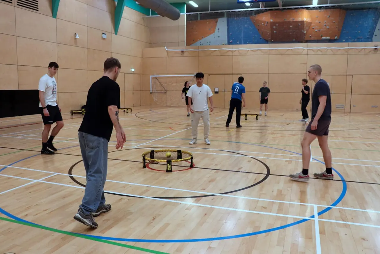 Group of young adults playing footgolf in an indoor sports hall with wooden floor and climbing wall visible in background.