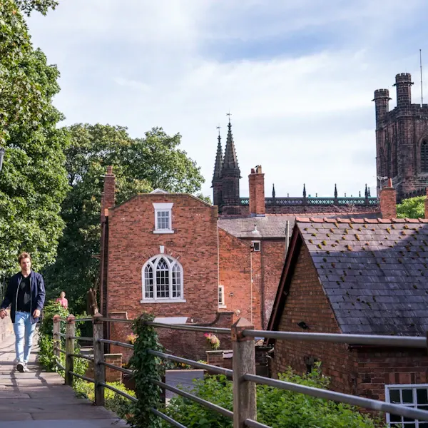 A person walks along a wooden-fenced pathway past red-brick buildings with ivy, with a historic church and spires visible in the background.