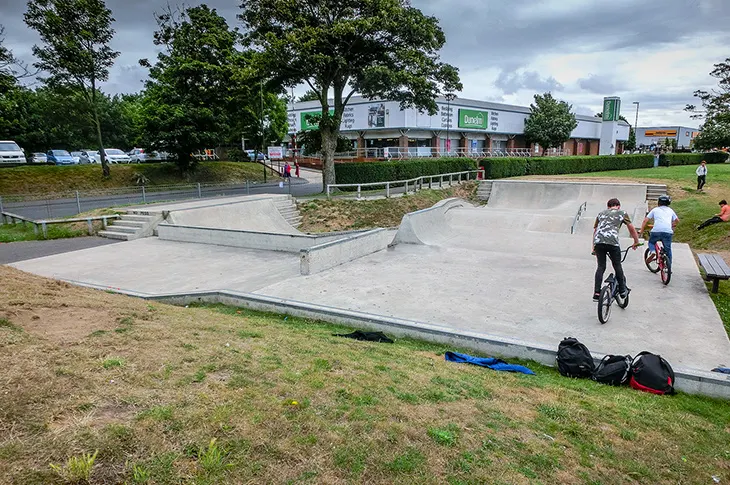 Young people riding and standing with bikes at a skate park with concrete ramps and features.