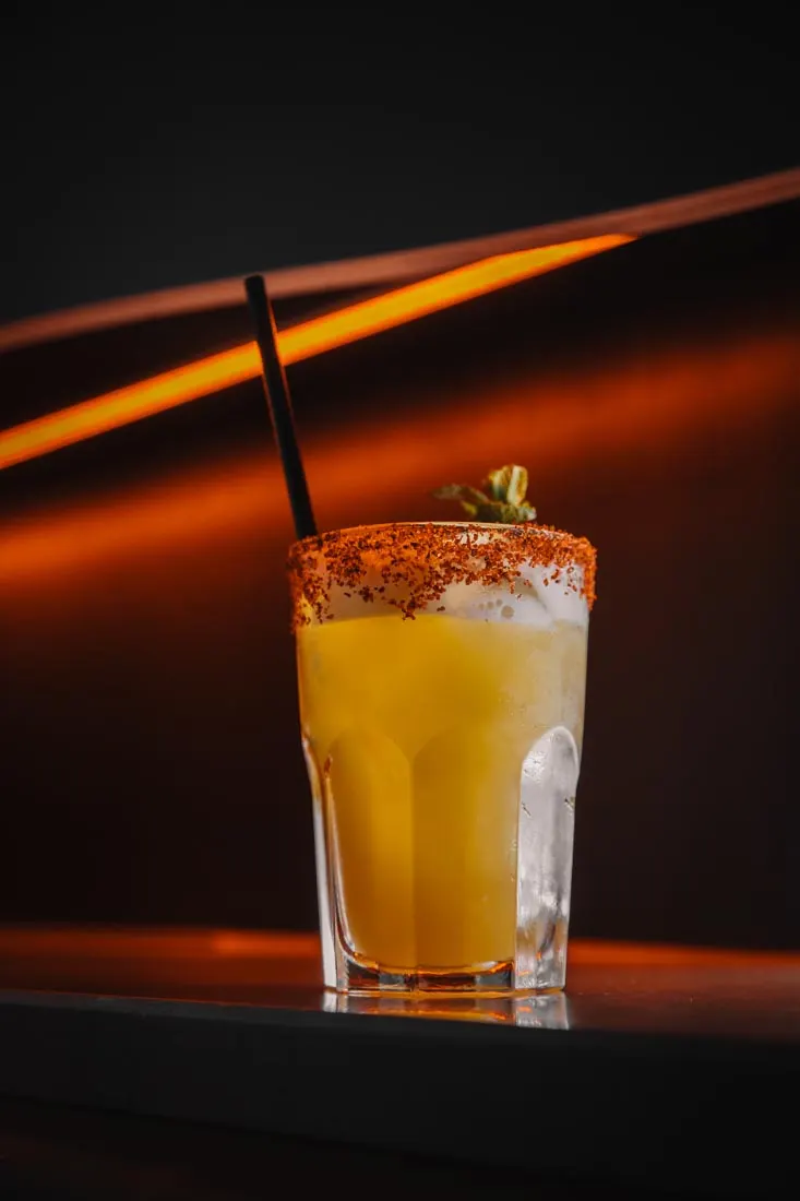 A chilled cocktail with amber liquid, spiced rim, and straw against moody orange and black lighting.