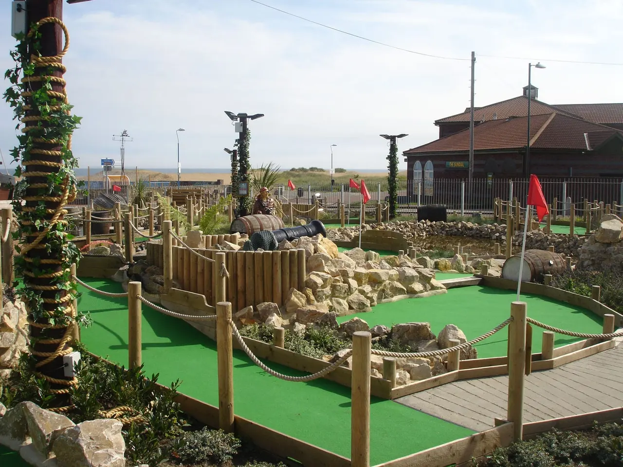 Mini golf course with bright green fairways, wooden railings, rock features, and red flags, beside a brick building overlooking coastal landscape.