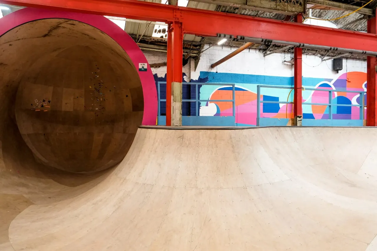 Indoor skate park with a large wooden barrel tunnel on the left and colourful murals on the walls under red steel beams.
