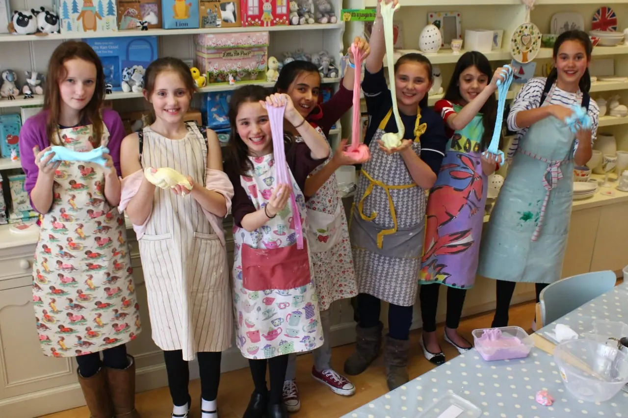 Children wearing handmade aprons display decorated ceramic pieces in a craft studio with shelves of pottery and supplies behind them.