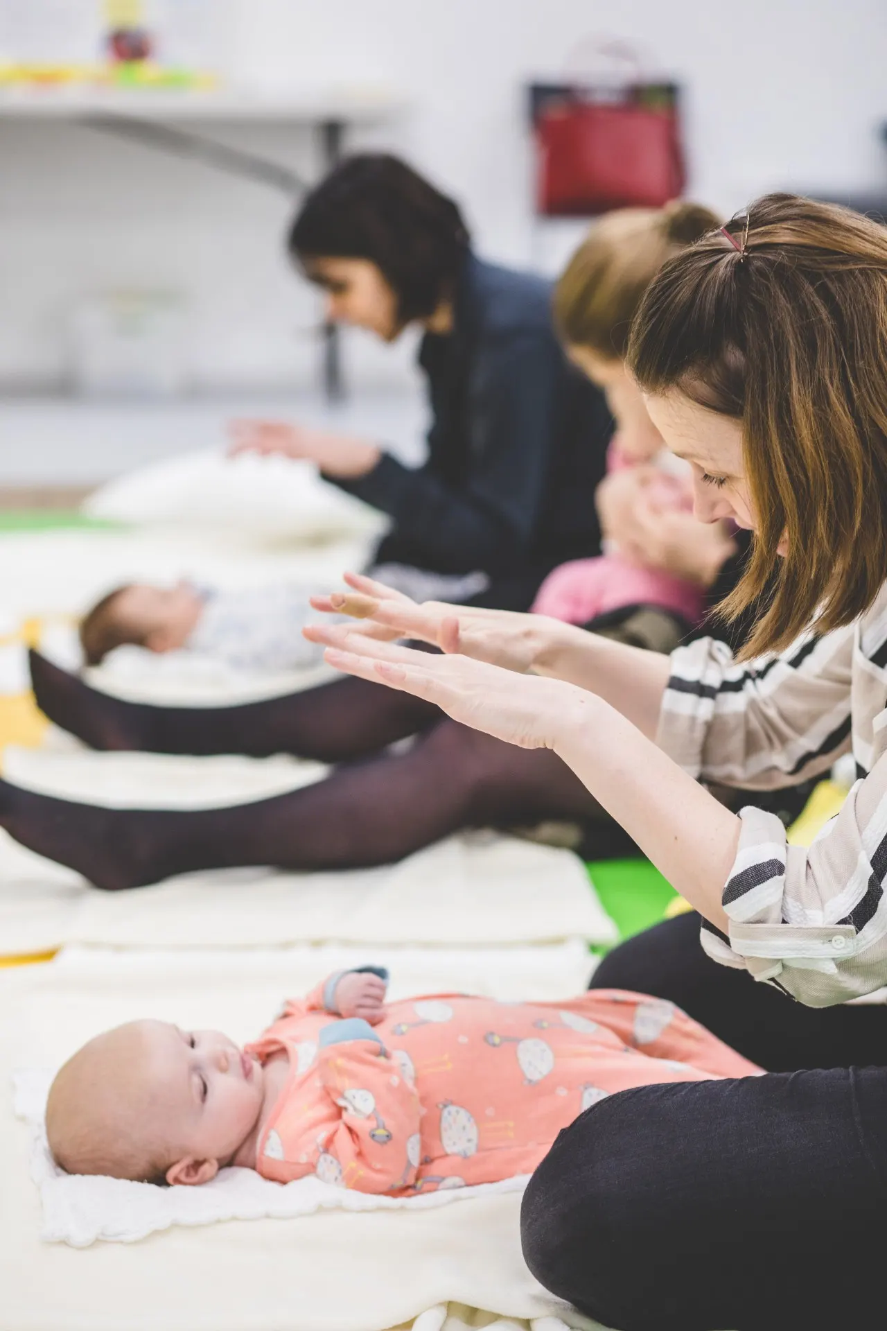 A woman guides a baby through a sensory activity whilst other parents and infants participate in a class session on mats.