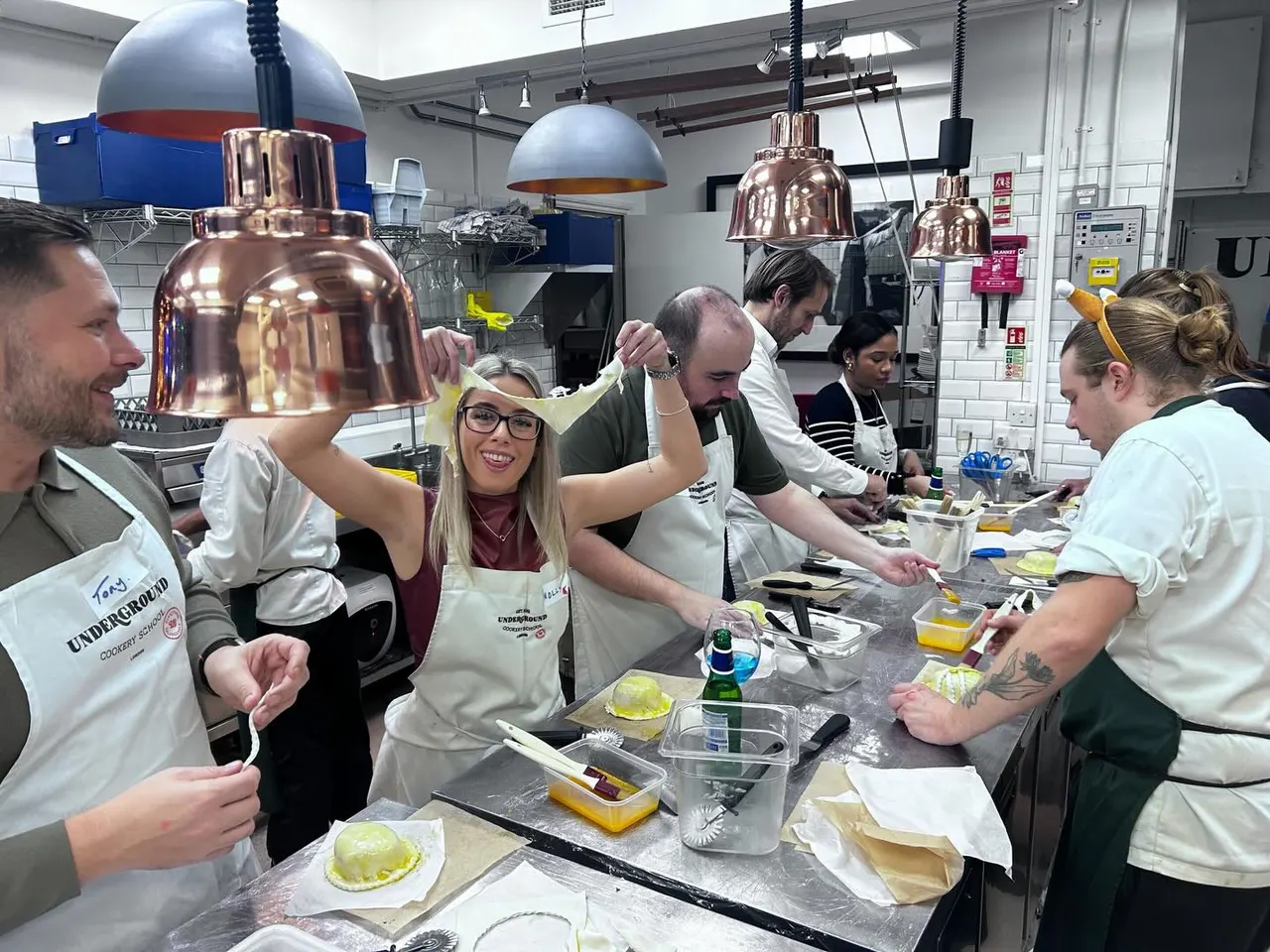 A group of people in aprons working together at a cooking class counter with copper heat lamps overhead.