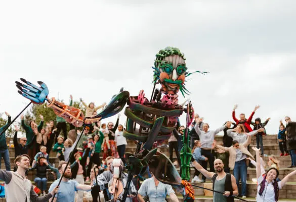 A large wooden effigy of a face surrounded by crowds of people with their arms raised in celebration outdoors.
