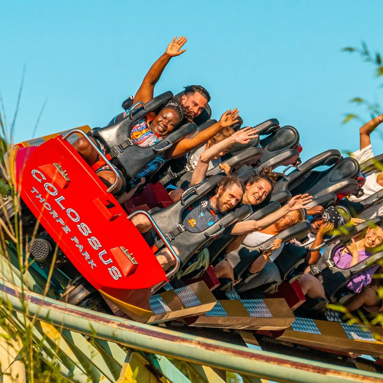 A group of people of various ages on a red roller coaster ride with arms raised in excitement against a clear blue sky.