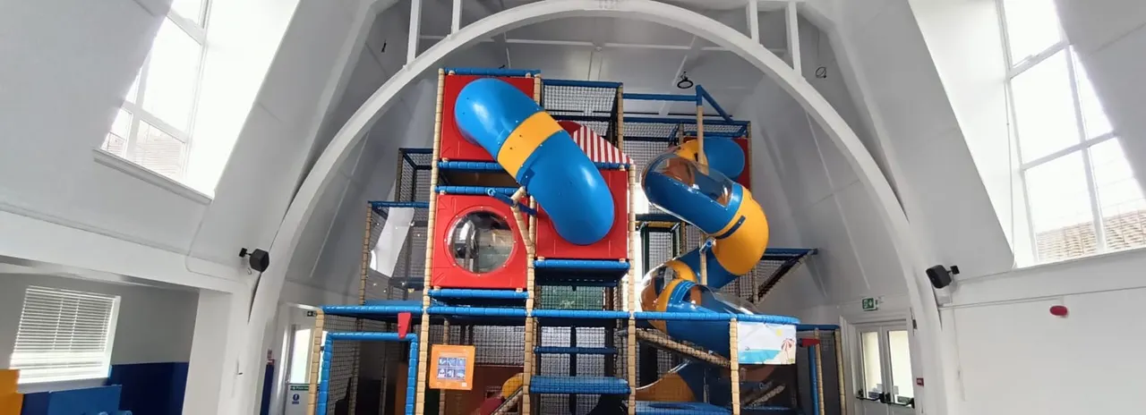Child climbing through a large indoor soft play structure with blue tunnels and slides in a bright white hall.