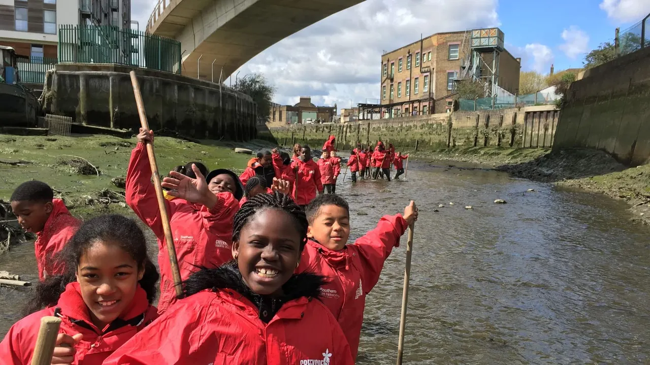 School children in red jackets holding poles whilst wading and playing in a shallow urban creek.