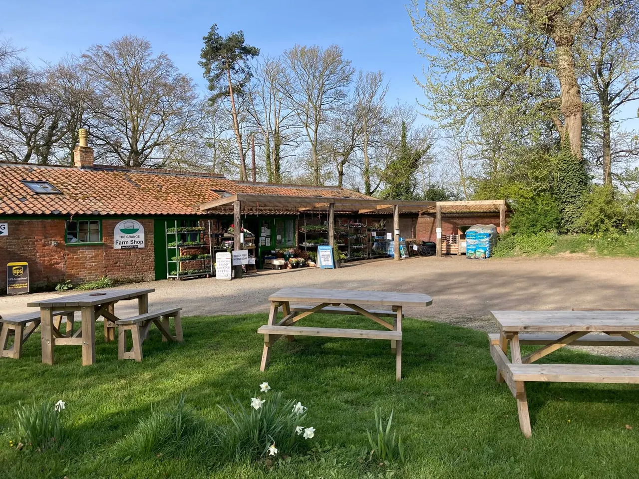 Picnic tables on grass in front of a farm shop building with trees in background on a sunny day.