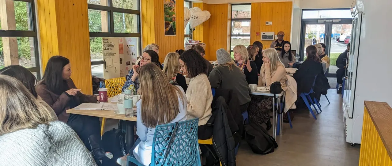 A group of adults gathered indoors at tables in a bright yellow-walled community space, socialising over refreshments.