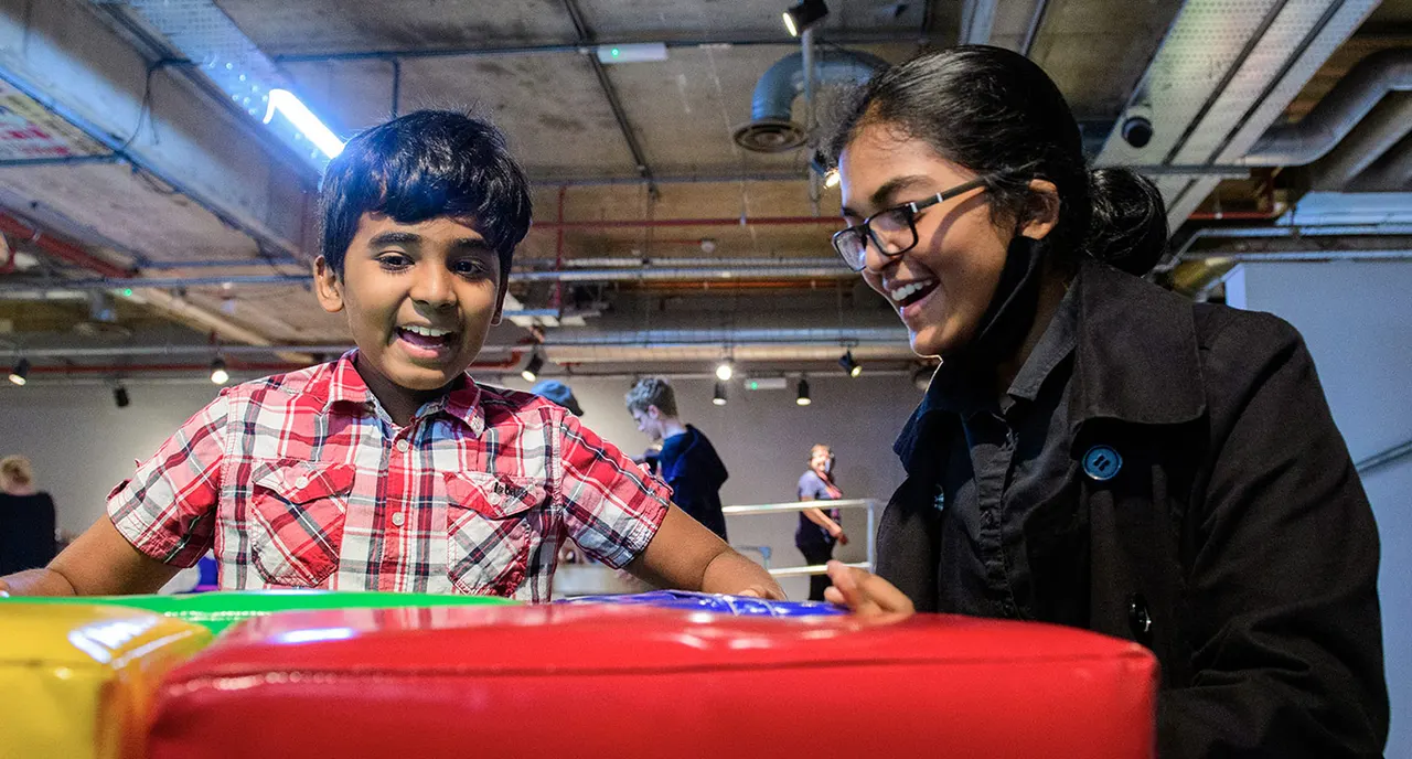 Two young people smiling while engaging with a colourful interactive display at an indoor science centre.