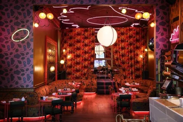 A sophisticated restaurant dining room with red and black décor, illuminated neon signs, and tables set for service.