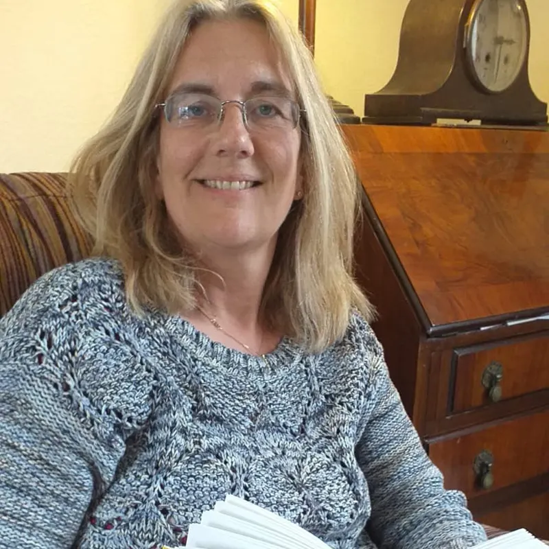 Woman with long grey hair and glasses sitting in striped armchair beside wooden furniture and clock.