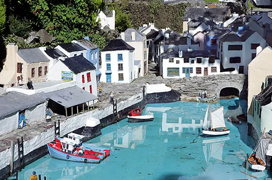 Miniature model village featuring tiny painted buildings, boats, and turquoise water in a seaside setting.