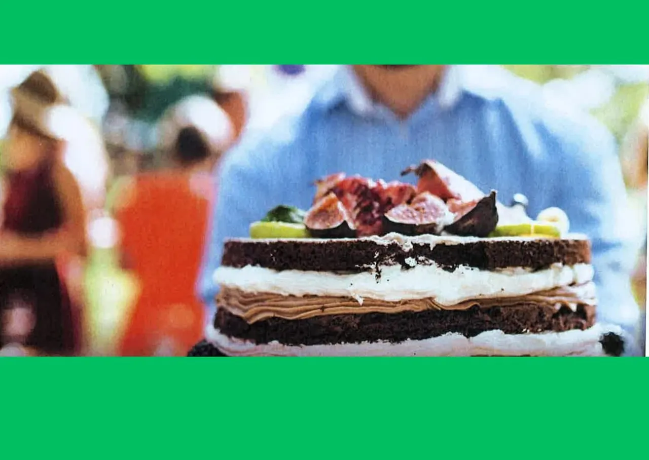 Layered chocolate cake with cream filling and fresh figs on top, displayed outdoors with people in background.