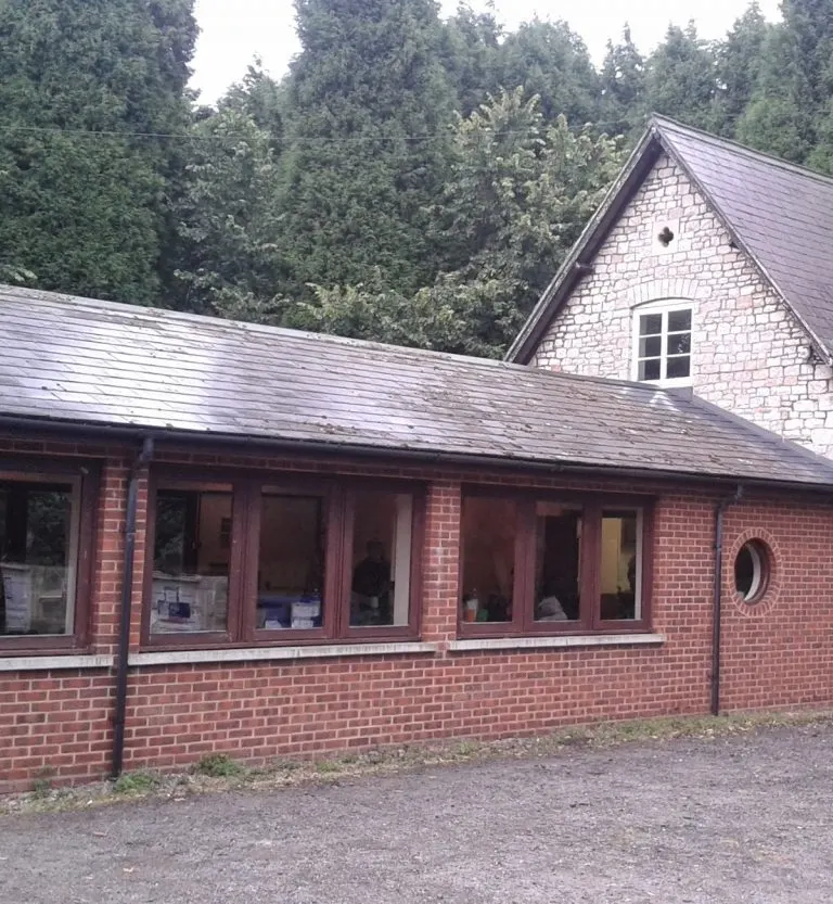 Red brick single-storey building with large windows and stone gable end, surrounded by dense woodland.