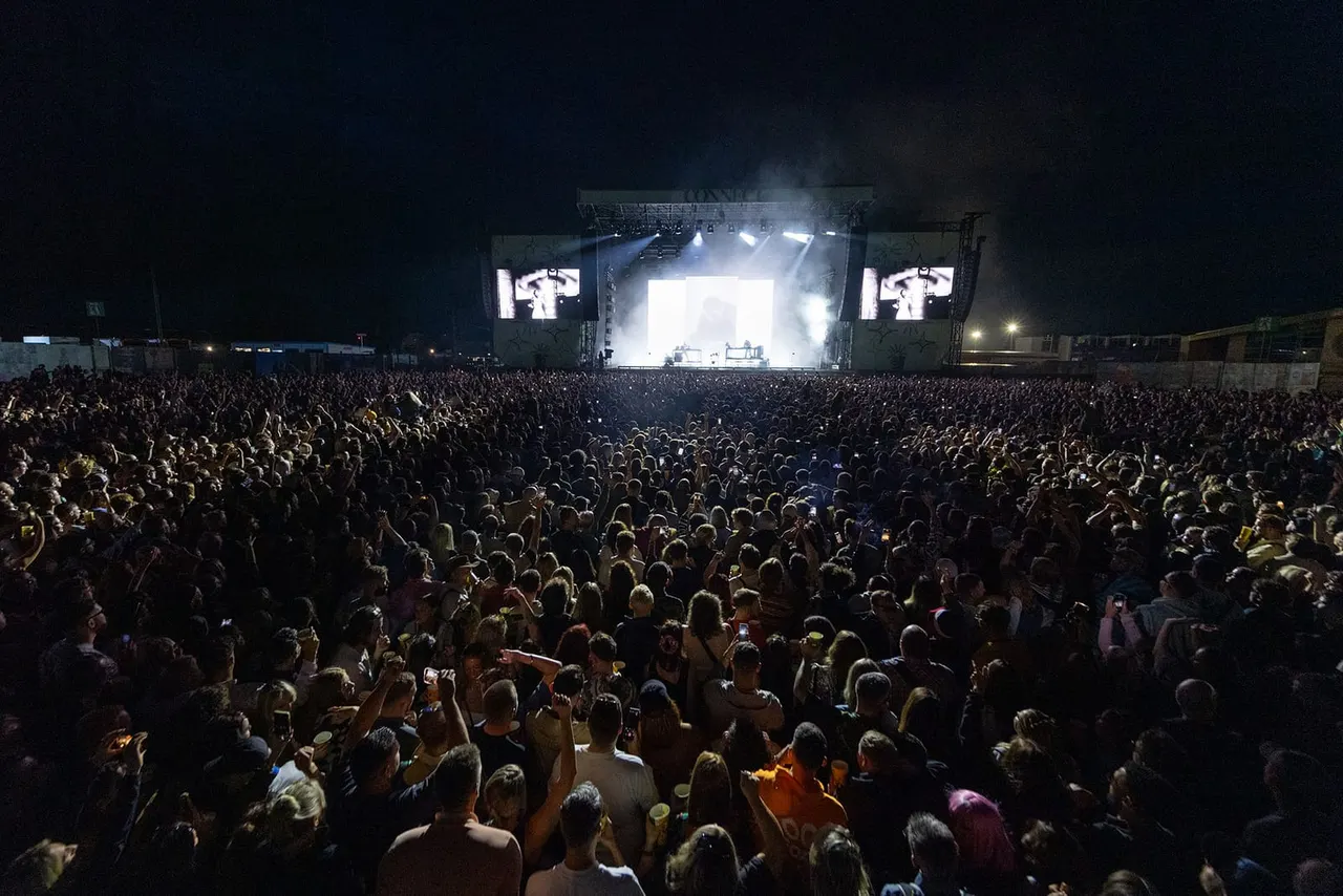 Large crowd of concert-goers at night watching a performer on a brightly lit stage with giant screens.