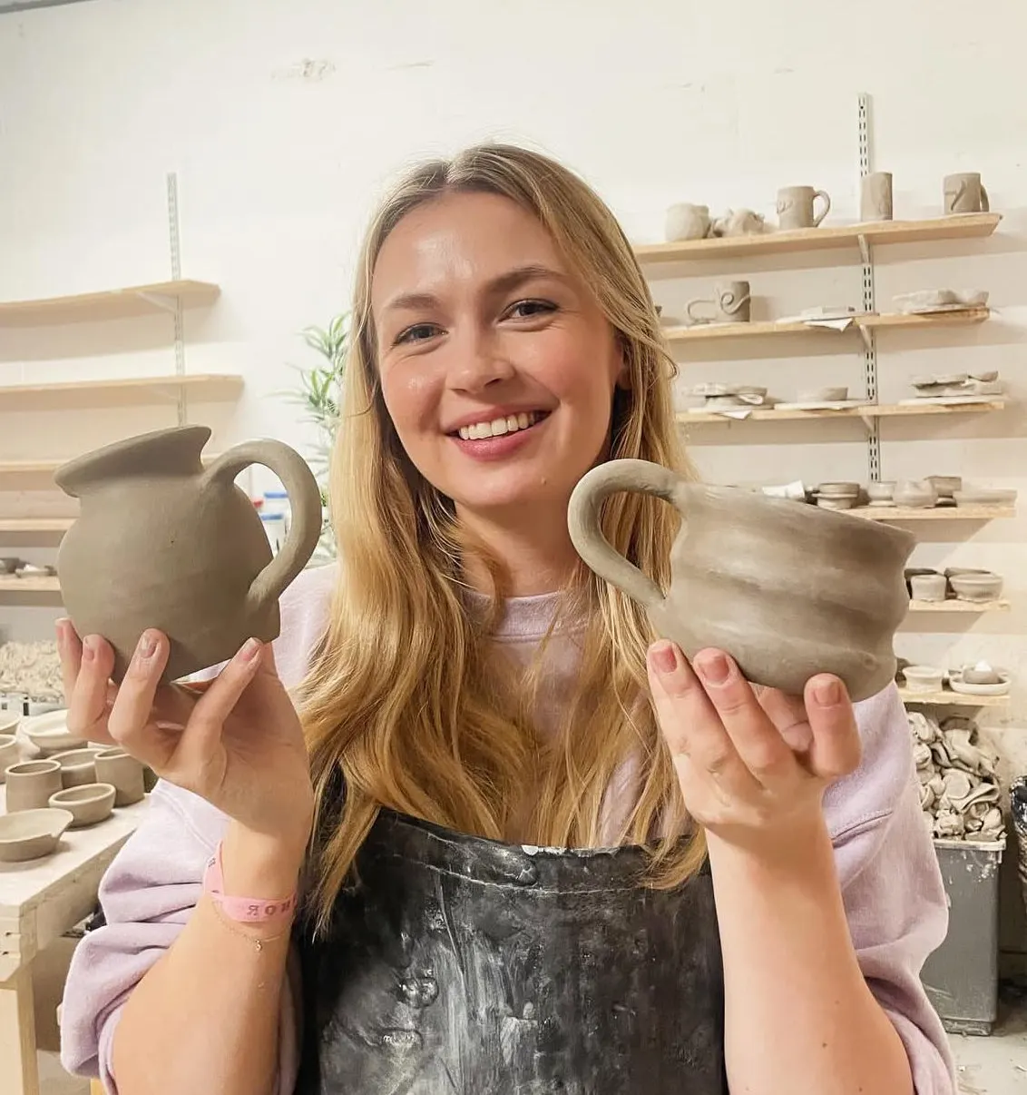 A young woman holding two clay vessels smiles at the camera in a pottery studio with shelves of ceramic pieces behind her.