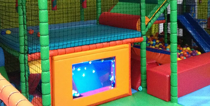 Colourful indoor play area with foam structures, netting, and a screen display within a soft play environment.