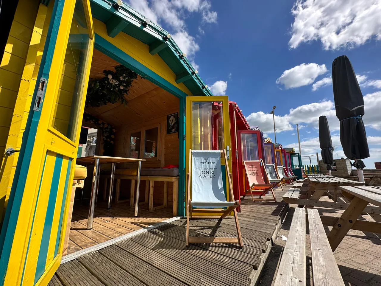 Colourful beach huts line a wooden deck under bright blue sky, with parasols and seating areas on a seaside promenade.
