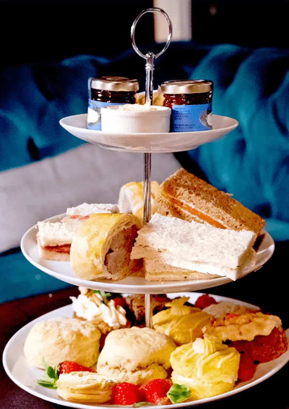 Three-tier stand displaying afternoon tea: scones with cream, sandwiches, pastries, and fresh strawberries, with condiments on top tier.