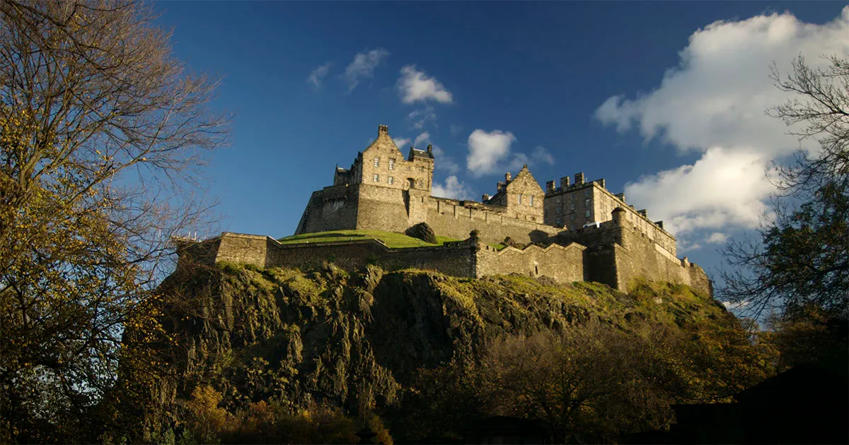 Stone castle perched on a rocky hilltop against a blue sky with white clouds and bare trees in the foreground.