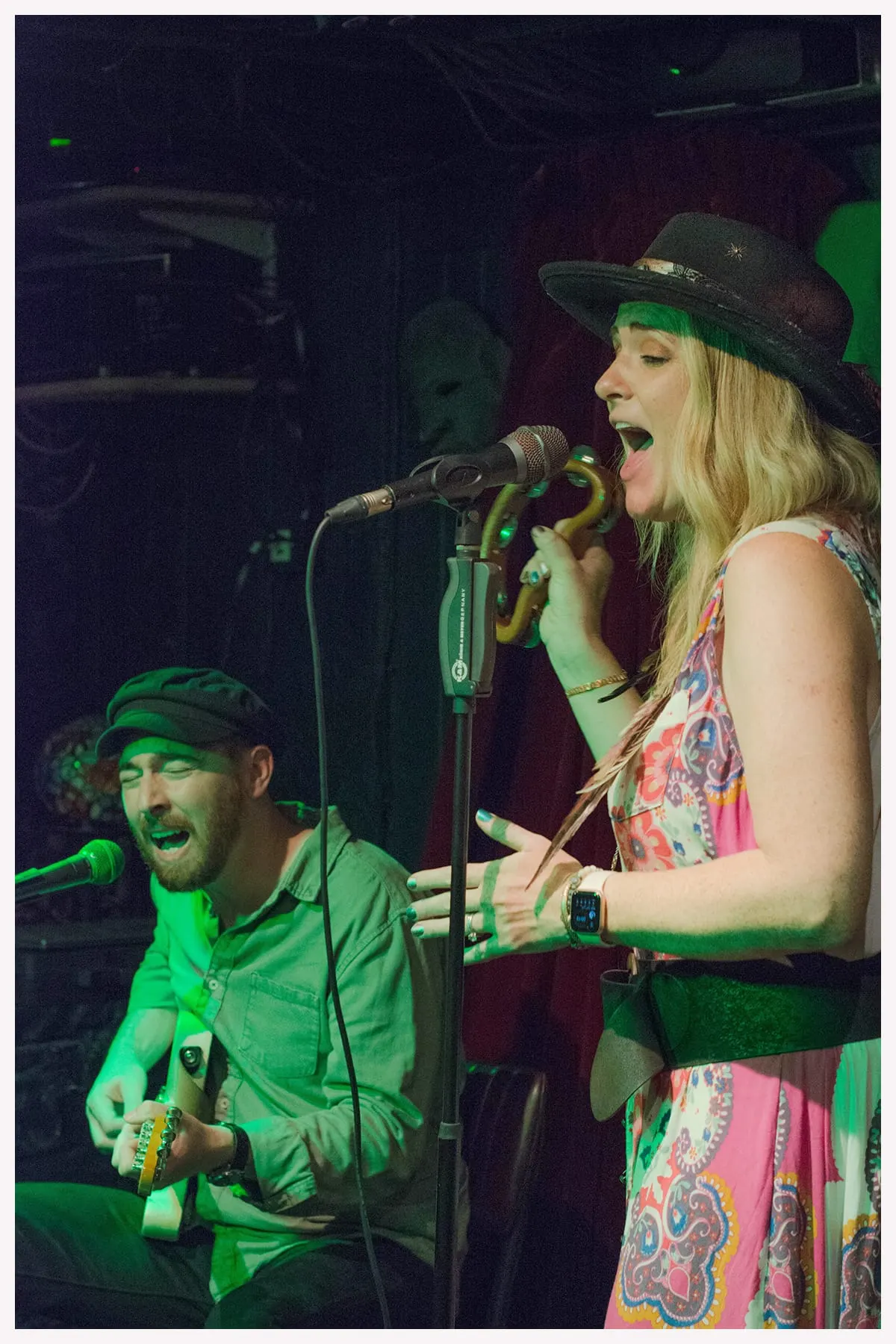 Two musicians performing on stage under green stage lighting, one seated with a guitar and one standing at the microphone wearing a black cowboy hat.