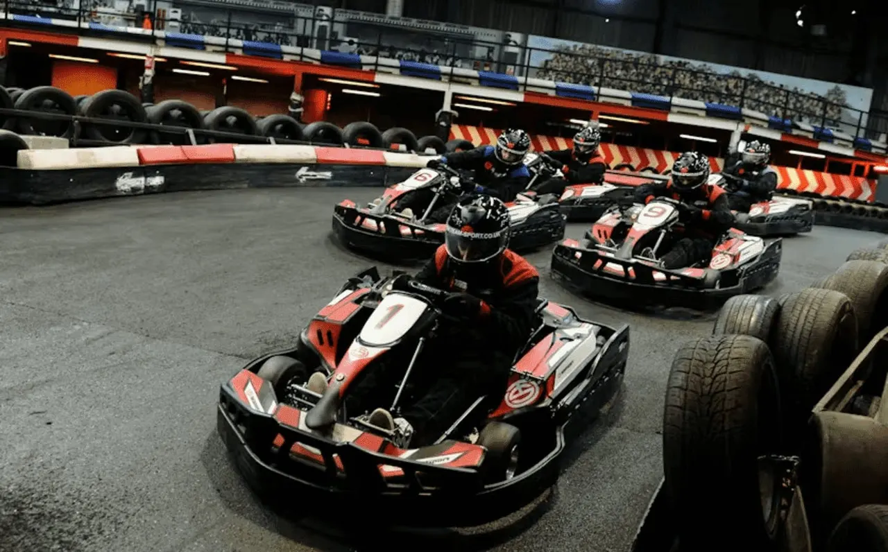 Go-kart racers in red and black karts racing around an indoor track with barriers and stadium seating.