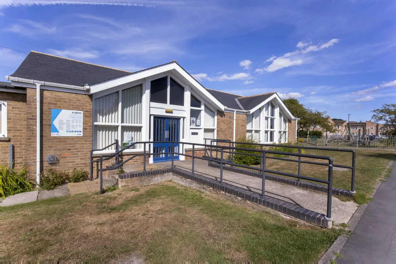 Modern brick and white leisure centre building with blue entrance doors and metal wheelchair ramp access, set in a grassy park area on a sunny day.