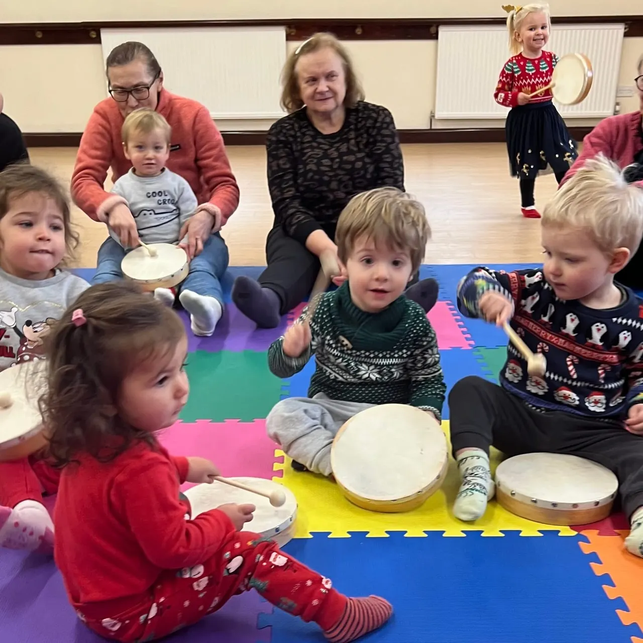 Young children and adults sit on a colourful foam mat playing drums and percussion instruments together in a music class.