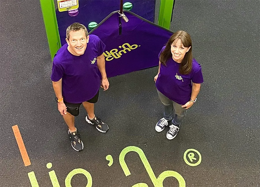 Two staff members in purple uniforms stand smiling at an indoor climbing wall facility with green and purple branding.