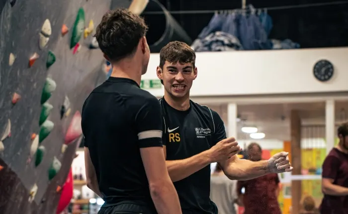 Two young men chat indoors at an indoor climbing wall with coloured holds.