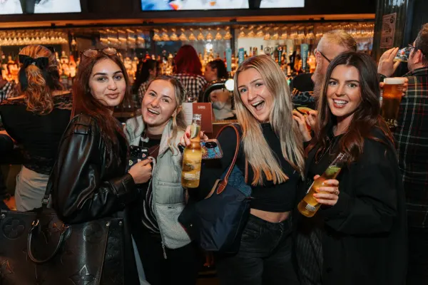 Four women holding drinks and smiling at the camera in a busy bar with illuminated shelving behind them.