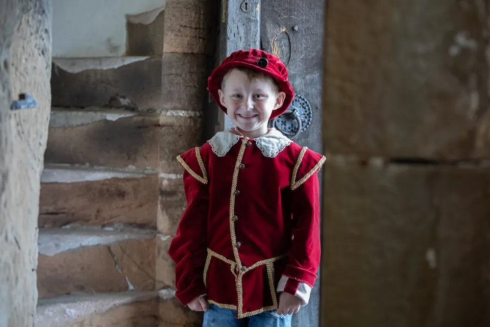 Young child in Tudor costume standing in stone passageway at historic manor house.
