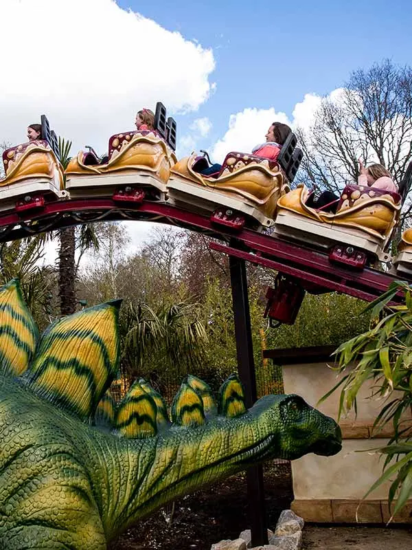 Visitors riding a golden dinosaur-themed roller coaster with a large green dinosaur sculpture in the foreground.