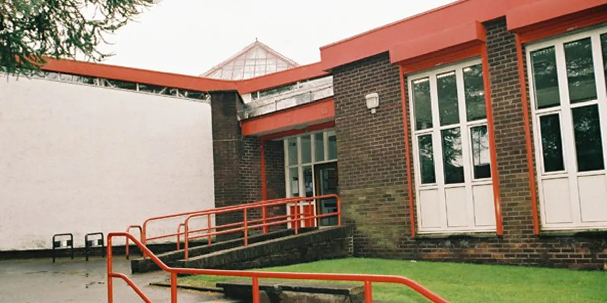 Exterior of a modern brick building with red metal railings and trim, featuring large windows and a glazed roof section, adjacent to a grassed area.