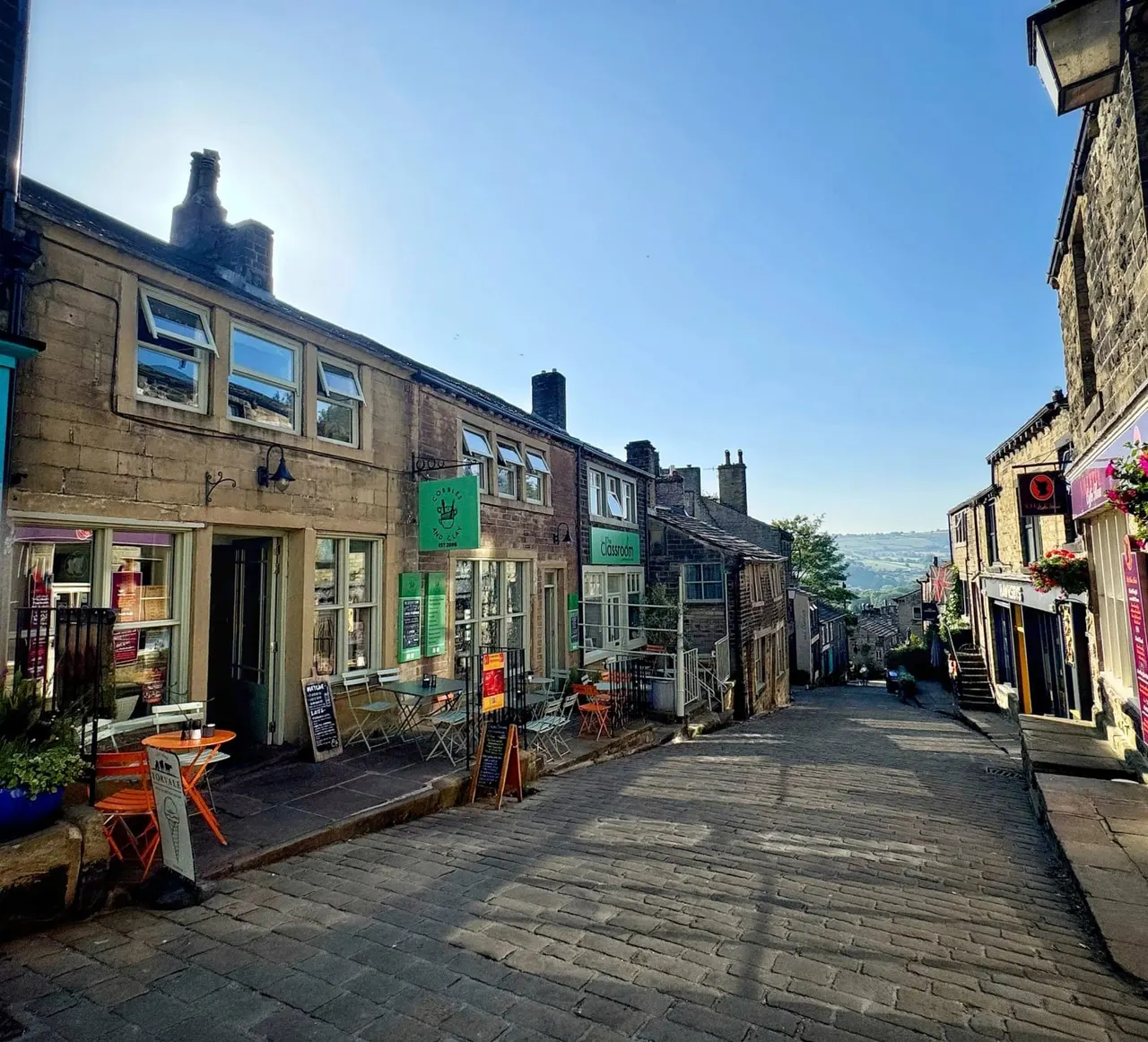 A sunny village street lined with stone buildings housing shops and cafés, with outdoor seating and hills visible in the distance.