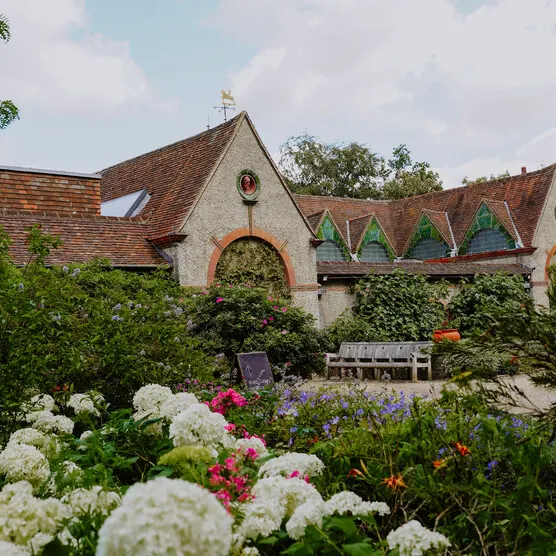 A historic stone building with arched windows surrounded by flowering gardens with white and pink blooms.