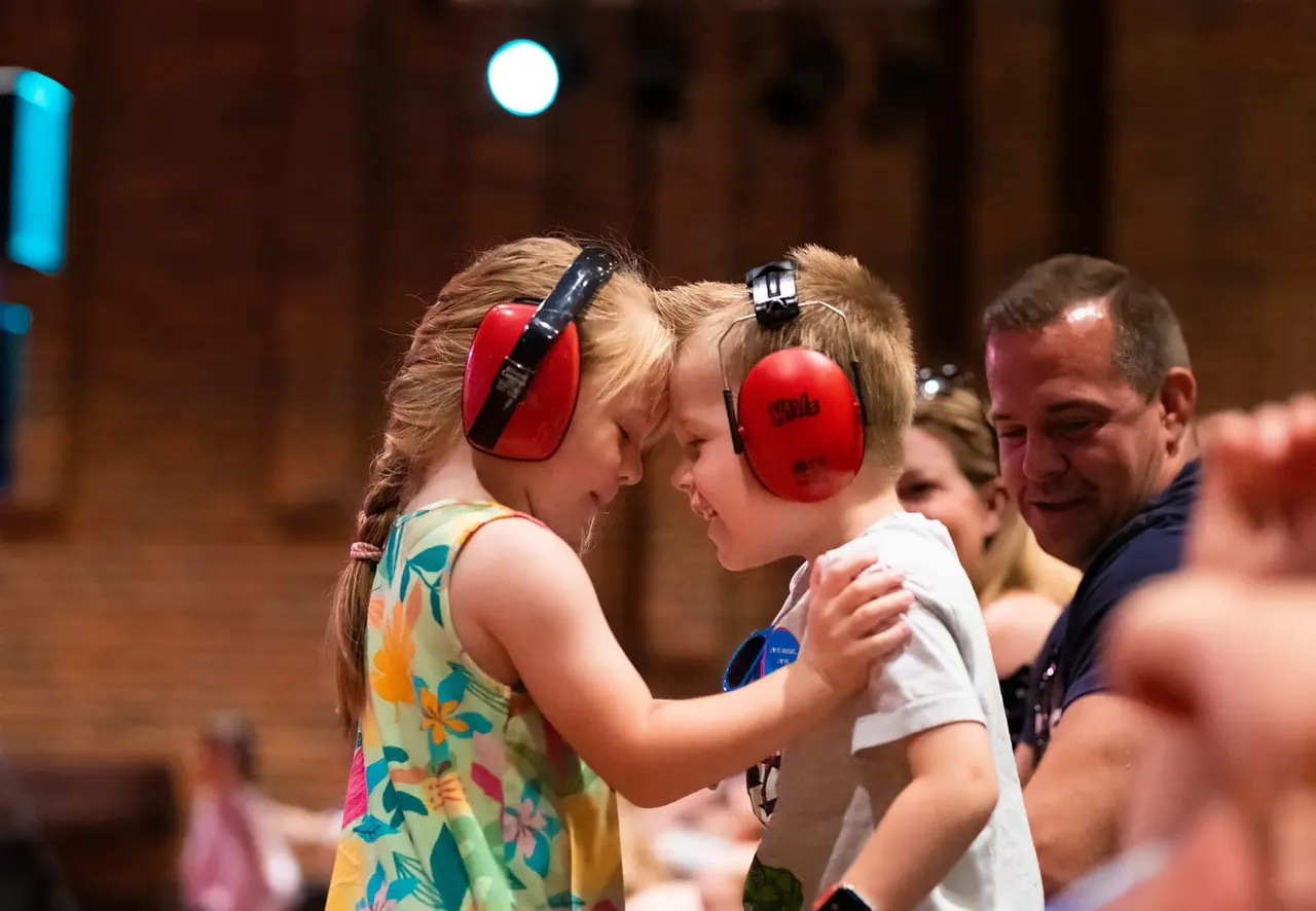 Two young children wearing red headphones face each other smiling in an indoor venue with adults nearby.