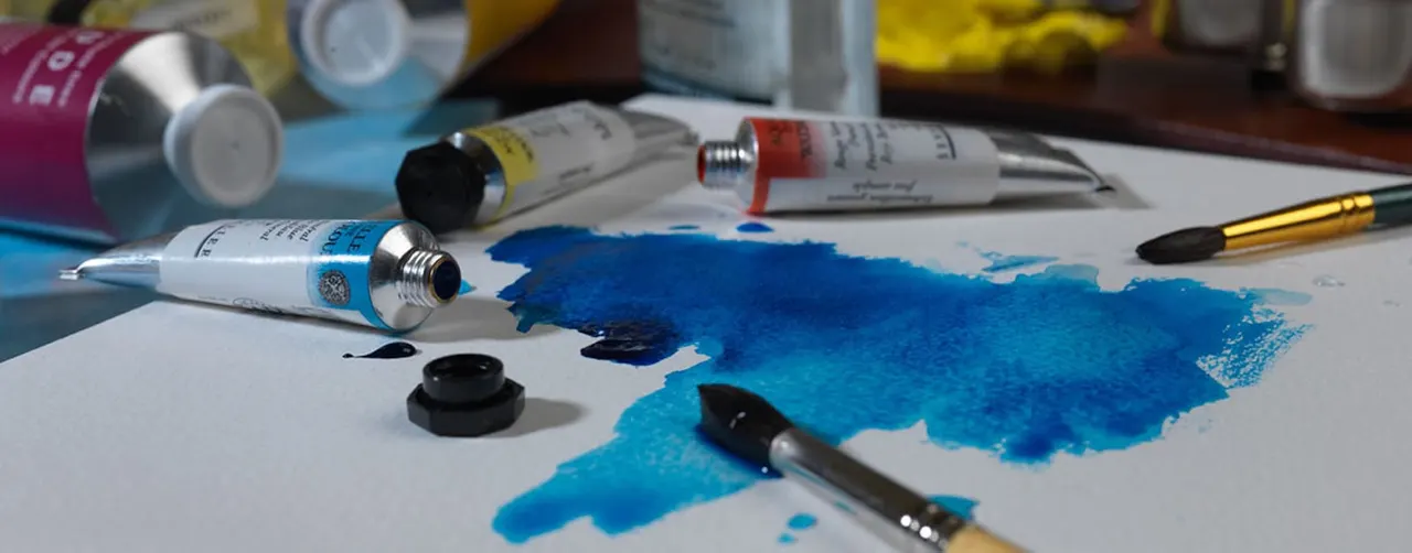 Art supplies including paint tubes, brushes, and a palette with blue watercolour paint on white paper.