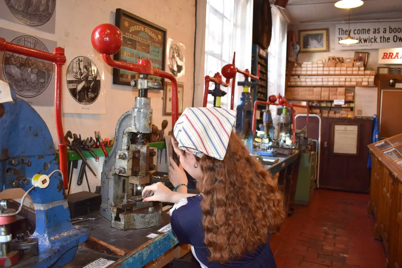 A visitor operates a vintage printing press in a museum workshop filled with historical equipment and memorabilia.