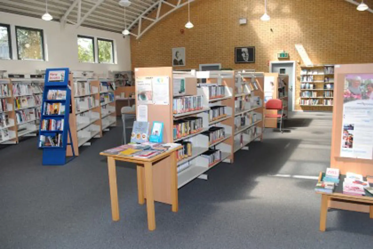 Interior of a library with wooden bookshelves, display tables, and a blue book stand under pendant lighting.
