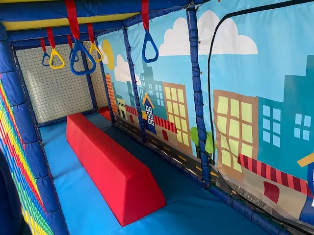 Colourful indoor play structure with suspended rings and trapeze bars above padded foam blocks, with cartoon cityscape wall graphics.