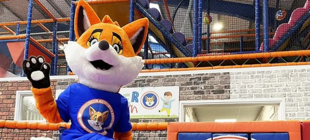 A smiling fox mascot in a blue shirt waves from an indoor play structure with orange and blue framework.