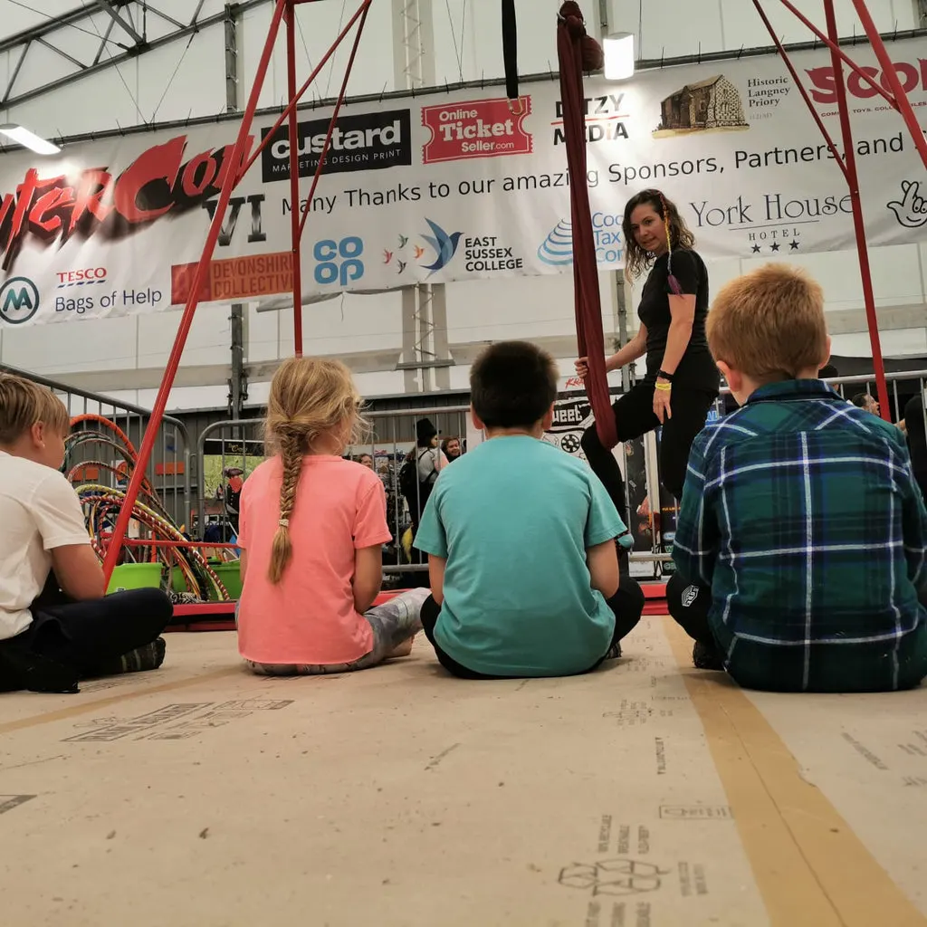 Children seated on the ground watching a circus performer on an elevated platform inside a large tent venue.