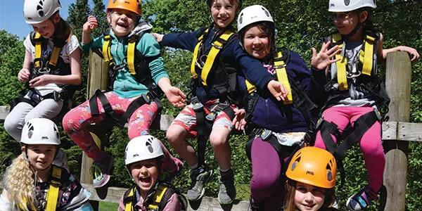 Group of children in safety helmets and harnesses sitting on wooden structure during outdoor activity session.